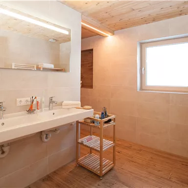 A modern bathroom with bright wood and an elegant tiled floor. The large mirror strip and windows provide plenty of light.