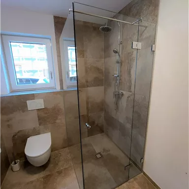 Ein modernes Badezimmer mit einer Glasdusche und einer Toilette. Die Wände sind mit neutralen, hellen Fliesen gestaltet.