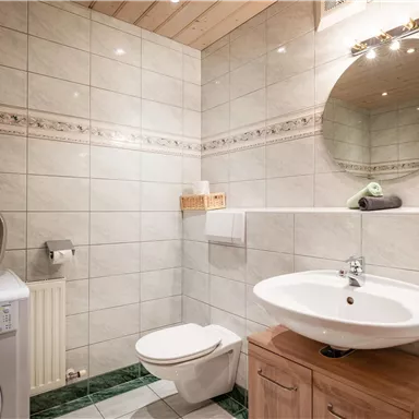 A modern bathroom with bright tiles and a round mirror. It features a sink, a toilet, and a washing machine.