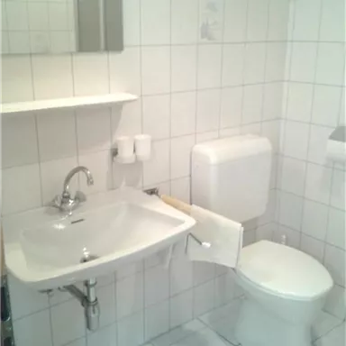 A small, clean bathroom with white tiles. There is a sink, a toilet, and a mirror.