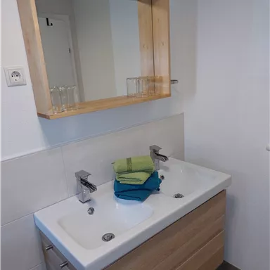 Ein modernes Badezimmer mit einem Doppelwaschbecken und einem Holzspiegel. Bunte Handtücher liegen ordentlich gestapelt auf dem Waschbecken.