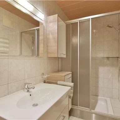 Ein modernes Badezimmer mit einer Duschkabine und einem Waschbecken. Die Wände sind hell gefliest und es gibt große Spiegel.