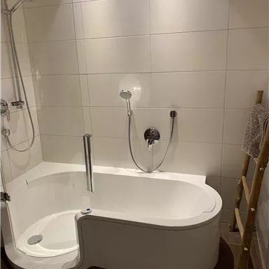 Ein modernes Badezimmer mit einer freistehenden Badewanne und Dusche. Die Wände sind hell gefliest und ein Holzregal ergänzt das Design.