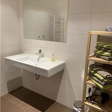 Ein modernes Badezimmer mit einem Waschbecken und einem großen Spiegel. Neben dem Waschbecken steht ein Regal mit sauberen Handtüchern.