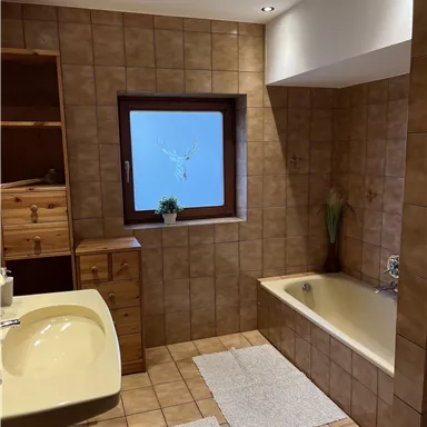 Ein gemütliches Badezimmer mit braunen Fliesen und einem Fenster. Es gibt eine Badewanne, ein Waschbecken und Holzregale.