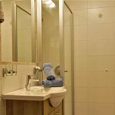 Ein helles Badezimmer mit Dusche, Waschbecken und Spiegel. Auf dem Waschtisch liegen zwei blaue Handtücher.