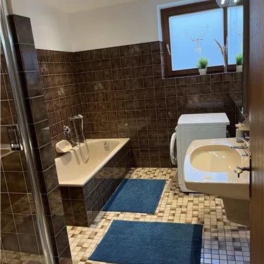 Ein modernes Badezimmer mit einer Badewanne, Dusche und Waschbecken. Die gefliesten Wände sind in Brauntönen gehalten, und es gibt blaue Teppiche auf dem Boden.