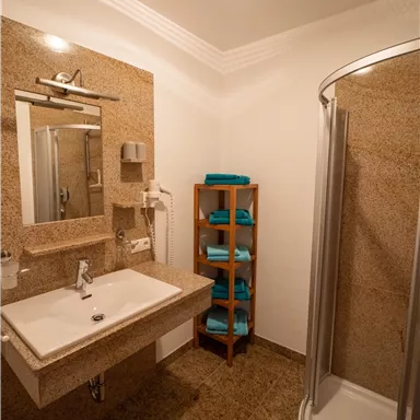 Ein modernes Badezimmer mit einer Dusche und einem Waschbecken. Auf einem Holzregal liegen frische, blau gefärbte Handtücher.