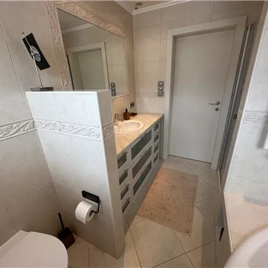 Ein modernes Badezimmer mit beigen Fliesen und einem großen Spiegel. Es verfügt über eine Waschbeckenanordnung und eine Tür, die zu einem weiteren Raum führt.