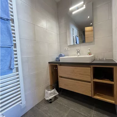 Ein modernes Badezimmer mit einer Holzkommode und einem Spiegel. Handtücher hängen an der Wand und der Boden ist aus dunklen Fliesen.