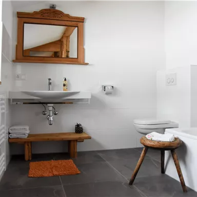 A modern bathroom with white walls and a wooden washbasin. There is a large mirror and a small orange rug.
