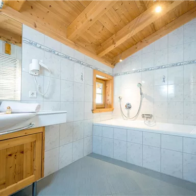 Ein modernes Badezimmer mit Holzelementen und hellen Fliesen. Die Badewanne und Dusche sind gut sichtbar, und viel Licht fällt durch die Fenster.