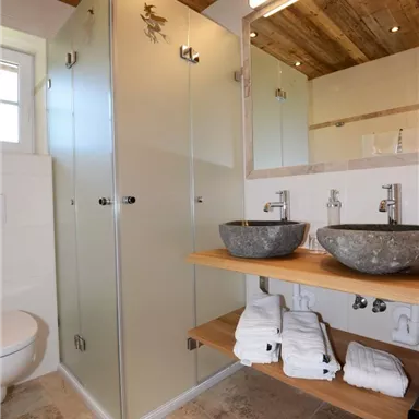 Ein modernes Badezimmer mit einer Glasdusche und eleganten Waschbecken aus Stein. Der Raum hat eine warme Holzdecke und helle Fliesen.