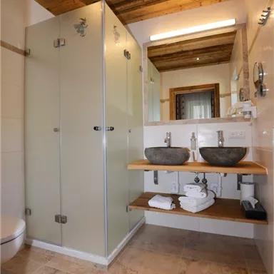 Ein modernes Badezimmer mit einer Dusche aus Glas und einem Holzregal. Zwei Waschbecken stehen auf dem Regal, zusammen mit Handtüchern und einem natürlichen Holzdecke.