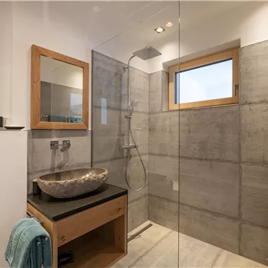 Ein modernes Badezimmer mit einer großzügigen Dusche und einem eleganten Waschbecken. Die Wände sind aus Beton, und es gibt viel natürliches Licht durch das Fenster.