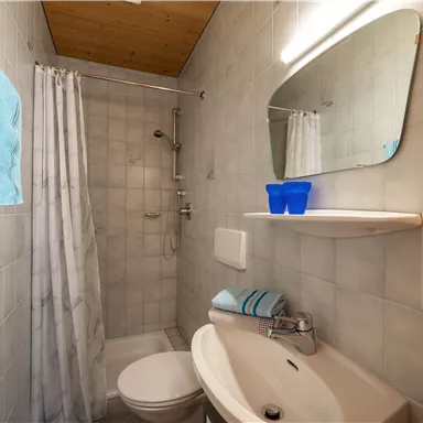 A small bathroom with a shower, sink, and toilet. The walls are tiled, and there are towels as well as a mirror shelf.