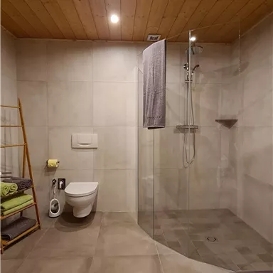 A modern bathroom with a spacious shower and a toilet. The walls are in neutral colors and there are fresh towels and a wooden towel holder.