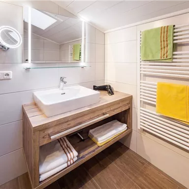 Ein modernes Bad mit Holzwaschtisch und einem großen Spiegel. Handtücher und farbige Badetücher hängen an einem Heizkörper.