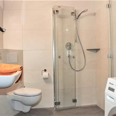 A modern bathroom with a shower cubicle and a washing machine. There is a sink with orange towels and a simple, light color scheme.