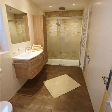 Ein modernes Badezimmer mit einer Dusche und elegantem Holzschrank. Der Boden ist braun gefliest und ein Handtuch liegt neben dem Waschbecken.