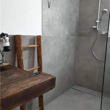 Ein modernes Badezimmer mit einer Duschkabine aus Glas und grauen Fliesen. In einer Ecke steht ein hölzerner Stuhl und ein Handtuchhalter.