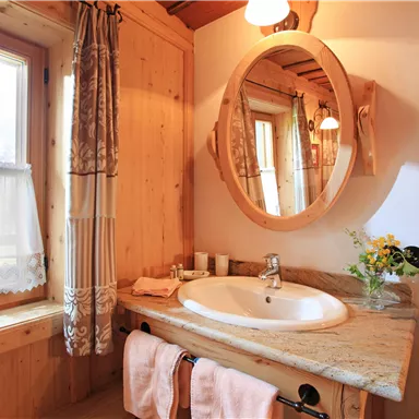 Ein gemütliches Badezimmer mit Holzverkleidung und einem großen Spiegel. Die Fenster bieten einen Blick nach draußen und es sind frische Blumen auf dem Waschbecken zu sehen.