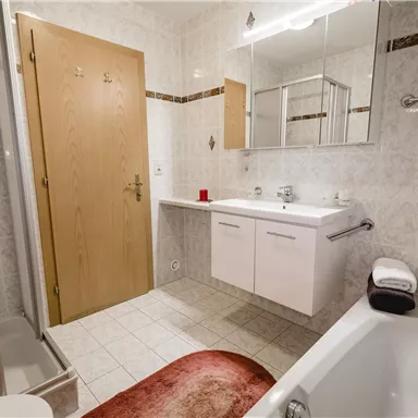 Ein modernes Badezimmer mit einer Dusche, einer Badewanne und einem Waschbecken. Die Wände sind hell gefliest und es gibt einen großen Spiegel.