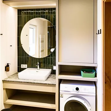 A modern bathroom with a round mirror and a sink. On the right, there is a washing machine in a simple cabinet.