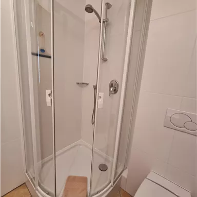 Eine moderne Duschkabine mit Glastüren in einem hellen Badezimmer. Der Boden ist gefliest und ein Handtuch liegt auf dem Boden.