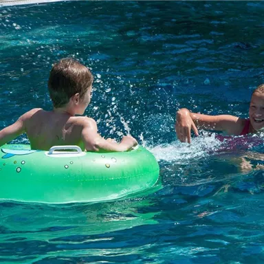 Ein Junge schwimmt auf einem aufblasbaren Ring im Wasser. Ein Mädchen spielt fröhlich neben ihm und spritzt Wasser.