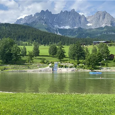 Ein ruhiger See umgeben von grünen Wiesen und Bäumen, mit beeindruckenden Bergen im Hintergrund. Der Himmel ist klar und blau.