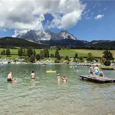 A clear lake surrounded by green meadows and mountains in the background. People are enjoying the water and the sun.