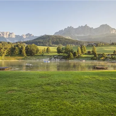 Eine ruhige Landschaft mit einem klaren Teich und grasbewachsenen Wiesen. Im Hintergrund erheben sich majestätische Berge unter einem blauen Himmel.