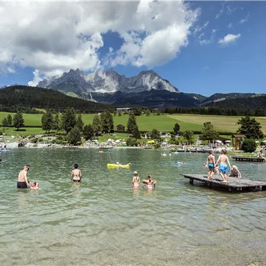 Ein klarer See umgeben von grünen Wiesen und Bergen. Menschen schwimmen und entspannen am Wasser.