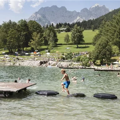 Ein Junge springt auf Schaumstoffinseln im Wasser. Im Hintergrund sind Schwimmer und eine grüne Wiese mit Bergen zu sehen.
