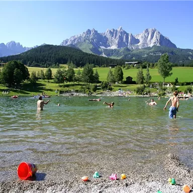 A beautiful swimming lake in the mountains with many people swimming in the water. In the background, green meadows and impressive mountain peaks can be seen.