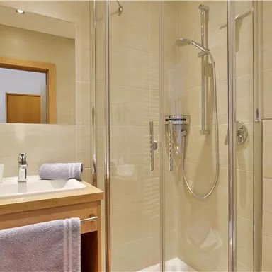 Ein modernes Badezimmer mit einer Dusche und einem Holzwaschtisch. Die Wände sind hell gefliest, und es sind Handtücher in sanften Farben vorhanden.