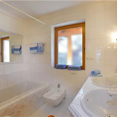 A bright bathroom with a bathtub and two sinks. The walls are painted in neutral colors, and there is a window allowing natural light.