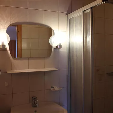 Ein Badezimmer mit einem Waschbecken, einem Spiegel und einer Duschkabine. Die Wände sind gefliest und es gibt einige Handtücher.