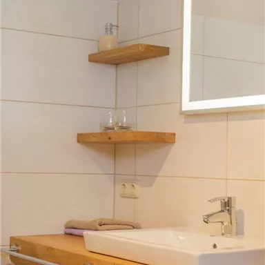 A modern bathroom with a wooden vanity and a mirror with lighting. Glasses and personal care products are placed on the shelves.