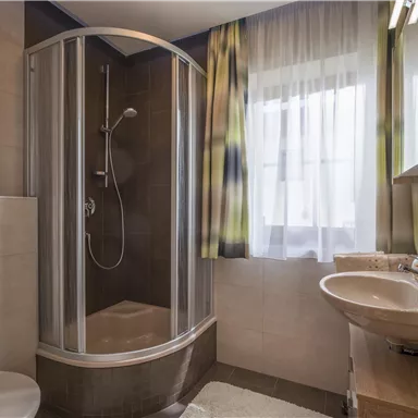 A modern bathroom with a shower and a sink. The large windows let in plenty of light, and the decoration is simple and elegant.