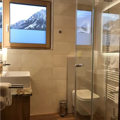 Ein modernes Badezimmer mit Dusche und Waschbecken. Durch das Fenster ist eine wunderschöne Berglandschaft sichtbar.