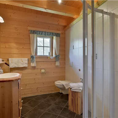 Ein gemütliches Badezimmer mit Holzwänden und einer Dusche. Es gibt ein Waschbecken sowie Handtücher und Fenster mit Vorhängen.