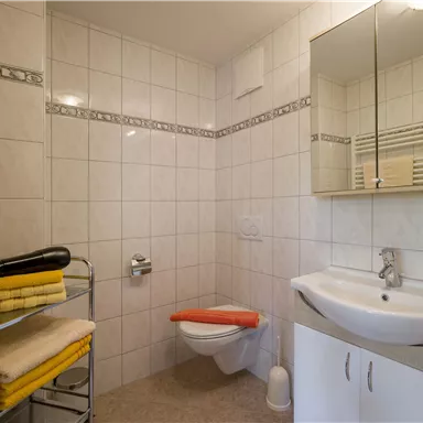 Ein helles Badezimmer mit weißen Fliesen und einem Waschbecken. Handtücher in gelb sind ordentlich auf einem Regal gestapelt.