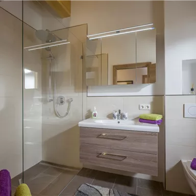 Ein modernes Badezimmer mit einer begehbaren Dusche und einem eleganten Waschbecken. Die Farben sind neutral, ergänzt durch lila Handtücher und grüne Dekorationselemente.