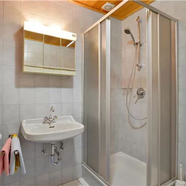 Ein modernes Badezimmer mit Dusche, Waschbecken und Spiegel. Die Wände sind hell gefliest und es gibt handtücher auf einem kleinen Regal.