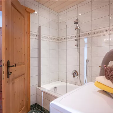 A modern bathroom with a bathtub and a shower. Towels are neatly placed on a white shelf, and the wooden door adds a rustic accent.