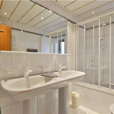 Ein modernes Badezimmer mit einem doppelten Waschbecken und einem Duschbereich. Die Wände sind gefliest und es gibt helle Holzoberflächen.