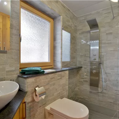 Ein modernes Badezimmer mit einer Dusche und einem Waschbecken. Die Wände sind mit grauen Steinfliesen verkleidet, und ein großes Fenster sorgt für Tageslicht.