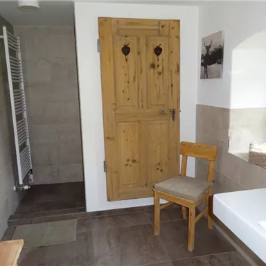 Ein modernes Badezimmer mit einer Holztür und einem Stuhl. Die Wände sind hell gehalten, und im Hintergrund ist ein Handtuchheizkörper sichtbar.